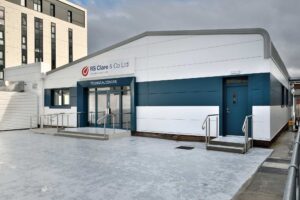 A modern building labeled "RS Clare & Co Ltd Technical Centre" with white and blue exterior panels. The building features a ramp and steps leading to glass entrance doors, with a railing along the walkway. A taller building is partially visible in the background.
