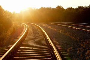A pair of railroad tracks curve gently to the left under a bright, setting sun. The sunlight casts a golden glow over the tracks and the surrounding landscape, with trees and vegetation lining both sides. Shadows stretch across the tracks, where ClareTech’s Curved Rail Grease ensures smooth passage.